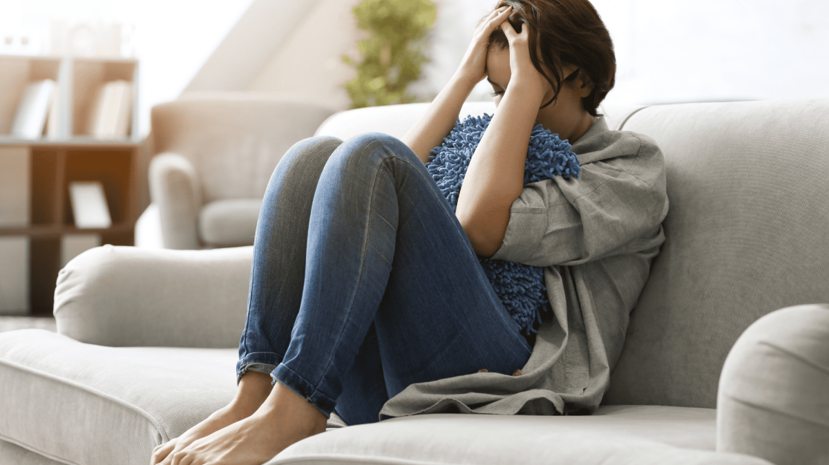 Frau sitzt mit angezogenen Beinen und Kopf in den Händen gestützt frustriert auf dem Sofa