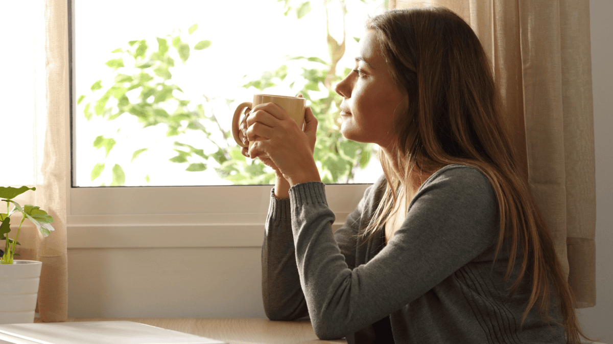 Frau mit Tasse in der Hand schaut nachdenklich aus dem Fenster.