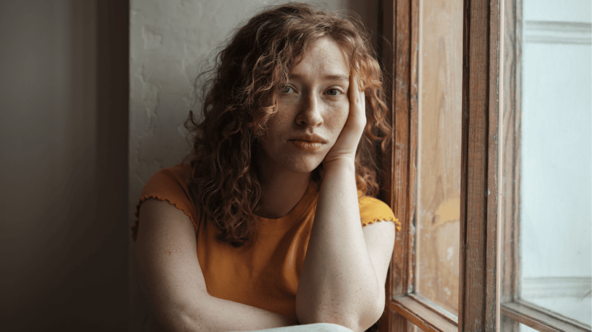 Frau mit roten Locken und gelbem T-Shirt sitzt am Fenster und schaut traurig in die Kamera