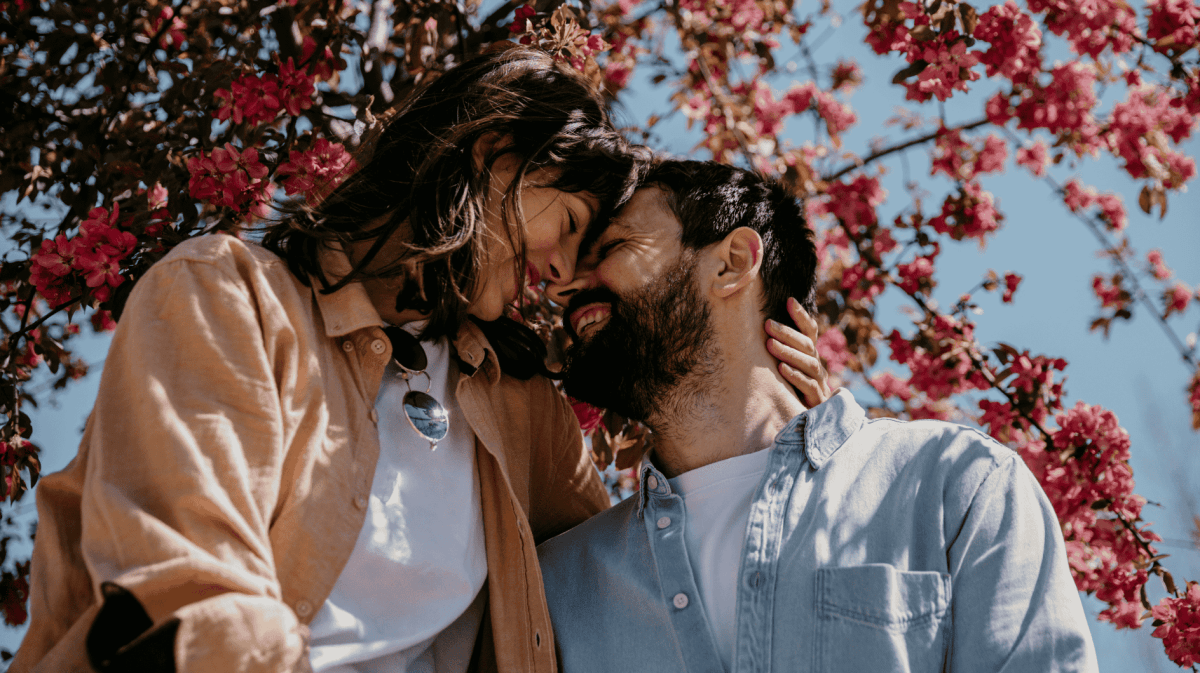Frau und Mann Stirn an Stirn schauen sich innig in die Augen umgeben von Kirschblüten und blauem Himmel bei Sonnenschein