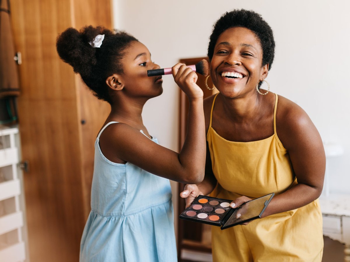 Tochter, ca. 10 Jahre alt, tr&auml;gt Rouge auf die Wange der Mutter auf. Beide stehen im Schlafzimmer.