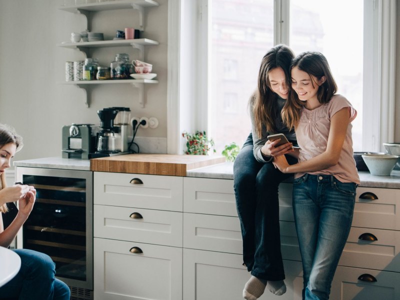 Drei Teenager-Mädchen in der Küche, schauen etwas auf ihren Smartphones nach.