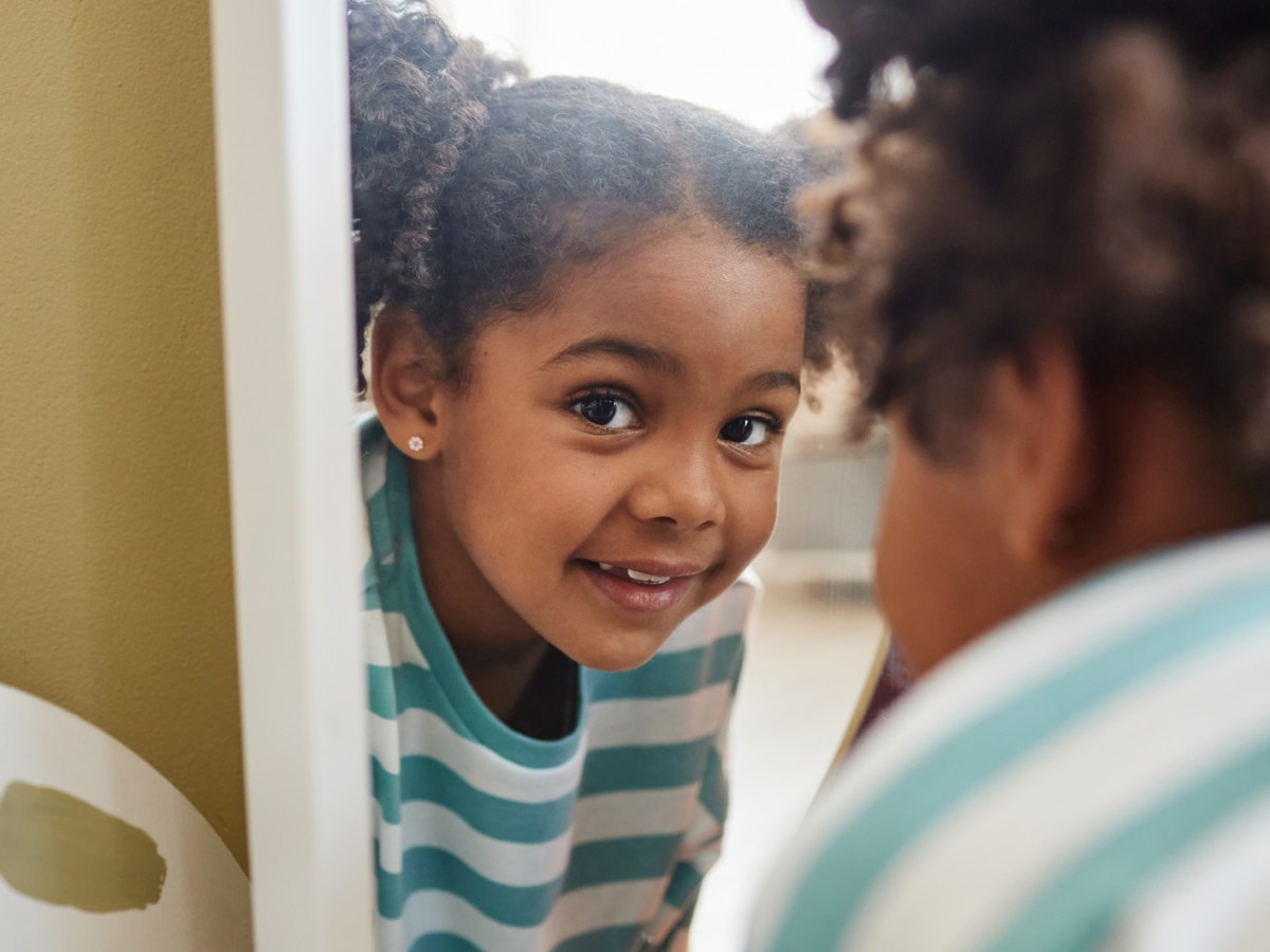 Nahaufnahme eines M&auml;dchens, das spielerisch &uuml;ber einen Spiegel in die Kamera lacht.