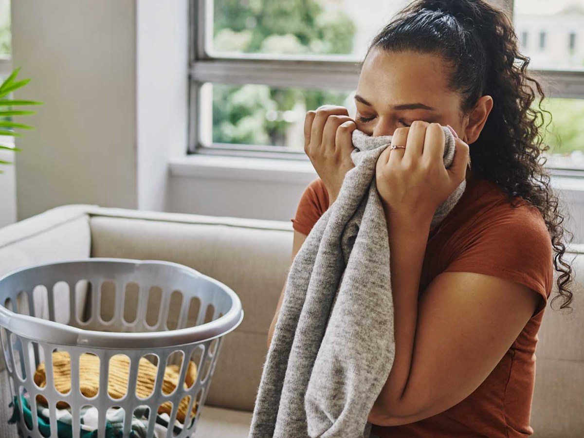 Junge Frau mit lockigem, dunklem Haar riecht l&auml;chelnd an grauem Pullover, neben ihr ein W&auml;schekorb auf beigem Sofa.