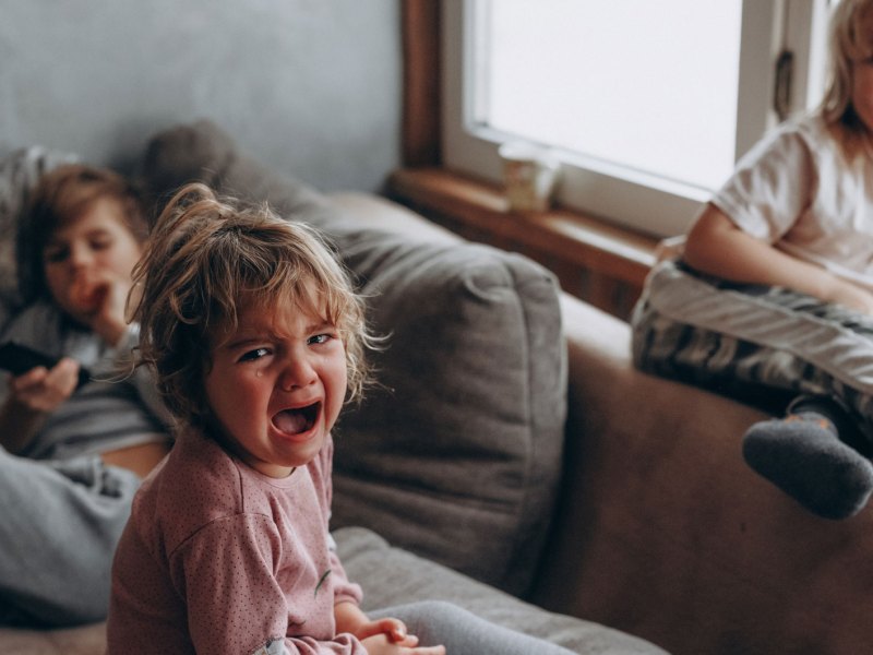 Drei Kinder auf dem Sofa. Das Große im Hintergrund hält die Fernbedienung, eines sitzt auf der Lehne und lacht und das dritte, vermeintlich jüngste, sitzt vorn auf dem Sofa und schreit und weint.