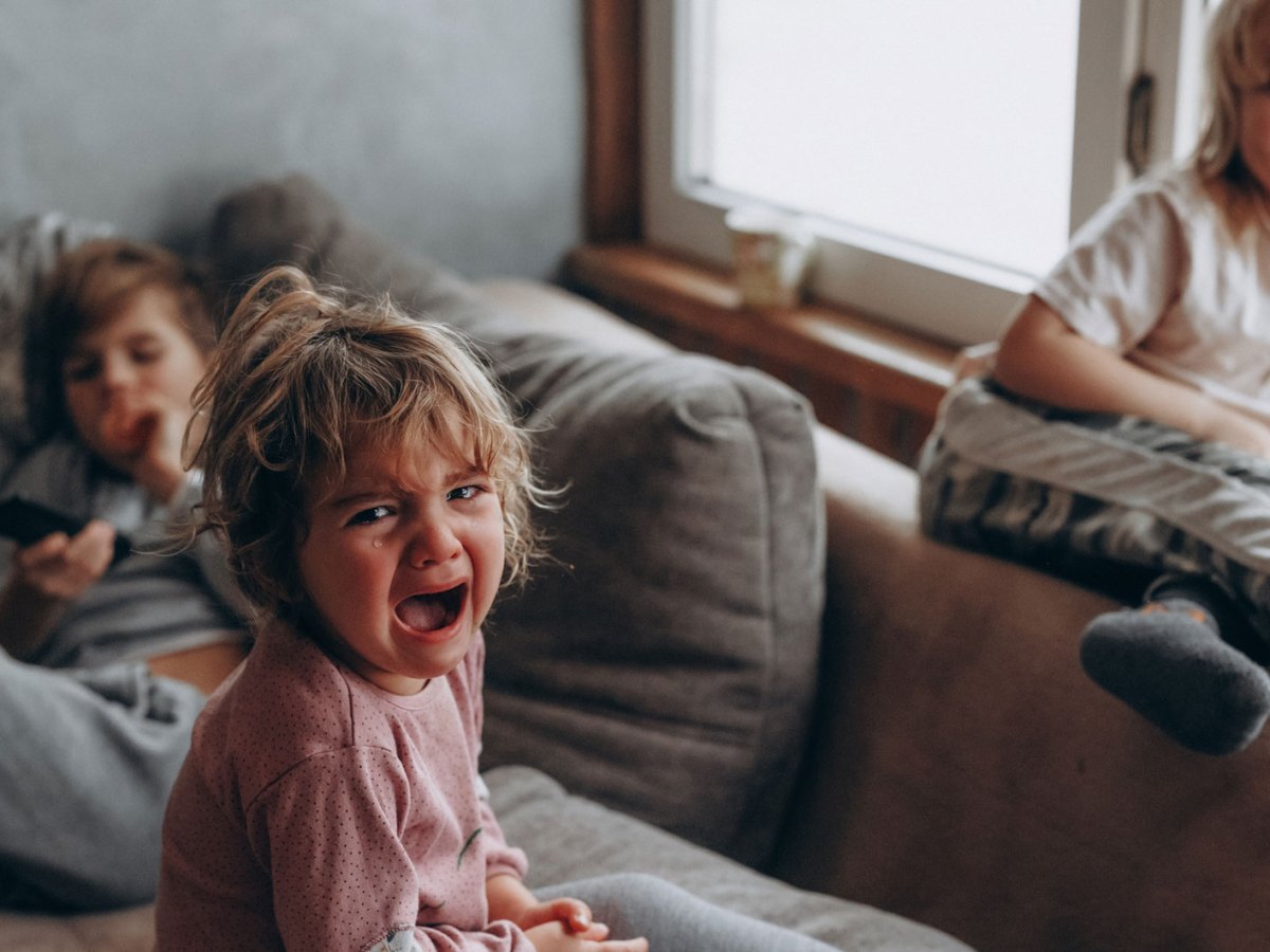 Drei Kinder auf dem Sofa. Das Gro&szlig;e im Hintergrund h&auml;lt die Fernbedienung, eines sitzt auf der Lehne und lacht und das dritte, vermeintlich j&uuml;ngste, sitzt vorn auf dem Sofa und schreit und weint.