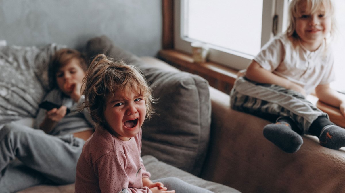 Drei Kinder auf dem Sofa. Das Große im Hintergrund hält die Fernbedienung, eines sitzt auf der Lehne und lacht und das dritte, vermeintlich jüngste, sitzt vorn auf dem Sofa und schreit und weint.
