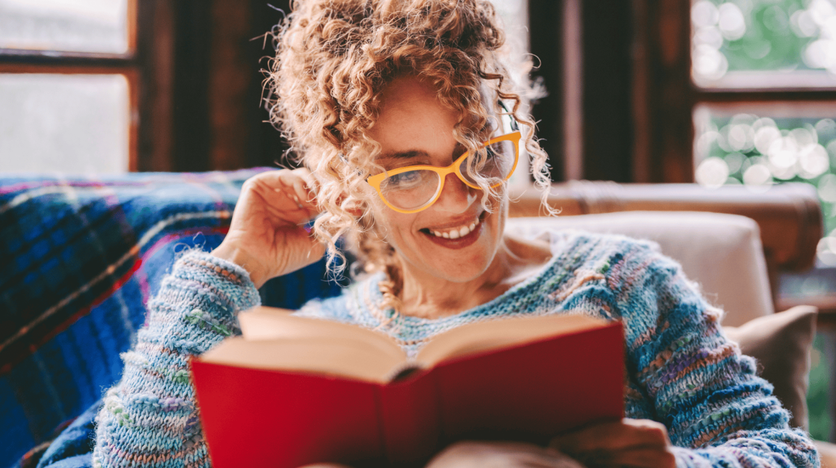Frau mit Locken sitzt auf Sofa und liest lächelnd ein Buch.