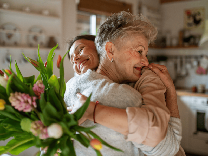 Junge Frau umarmt ihre Mutter und überreicht ihr Blumenstrauß.