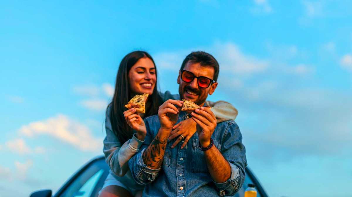 Paar sitzt auf einem Autodach und isst Pizza. Hinter ihnen strahlend blauer Himmel.