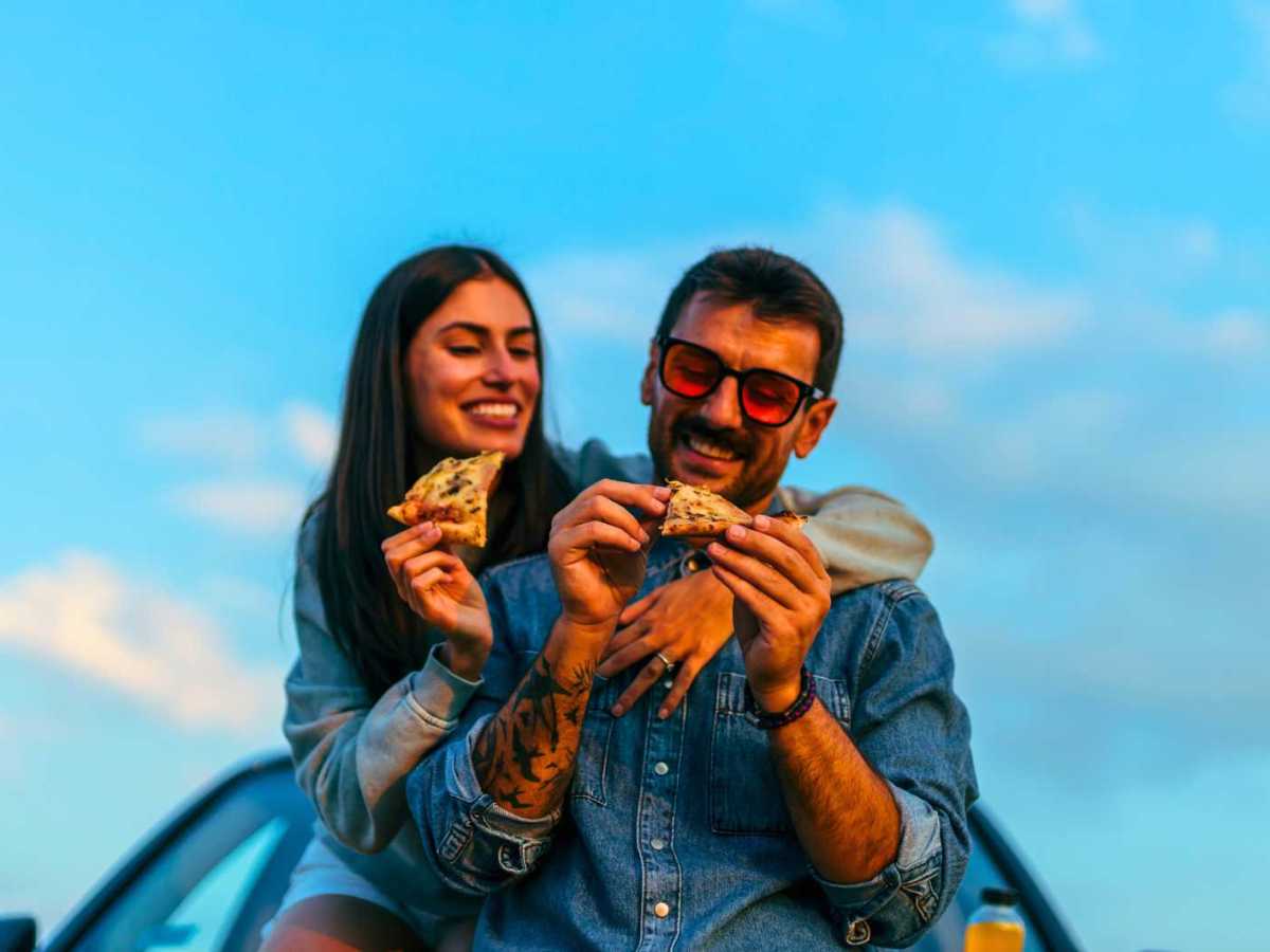 Paar sitzt auf einem Autodach und isst Pizza. Hinter ihnen strahlend blauer Himmel.