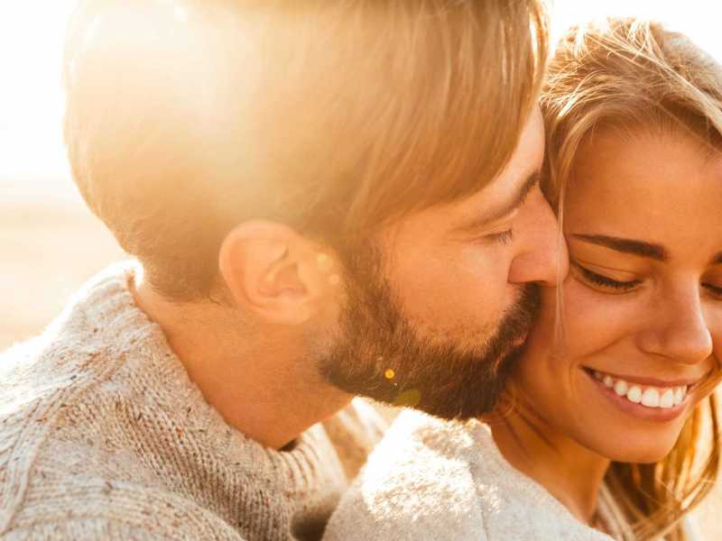 Mann umarmt seine Freundin und gibt ihr einen Kuss auf die Wange. Sie lächelt.