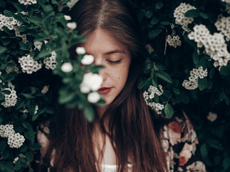 Brünette Frau steht vor Gebüsch mit Blumen.