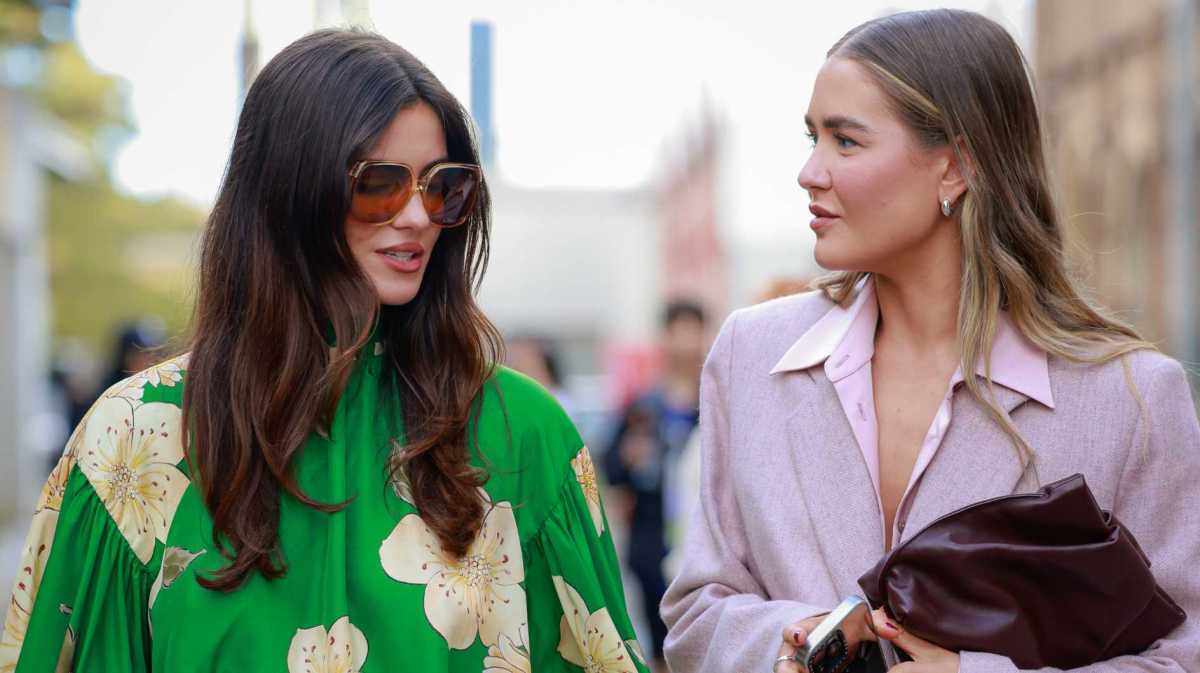 Frau trägt braune Sonnenbrille, grünes Oberteil mit Blumenmuster, gelben Minirock, schwarze Stiefel und eine Tasche. Zweite Frau trägt rosa Anzug.