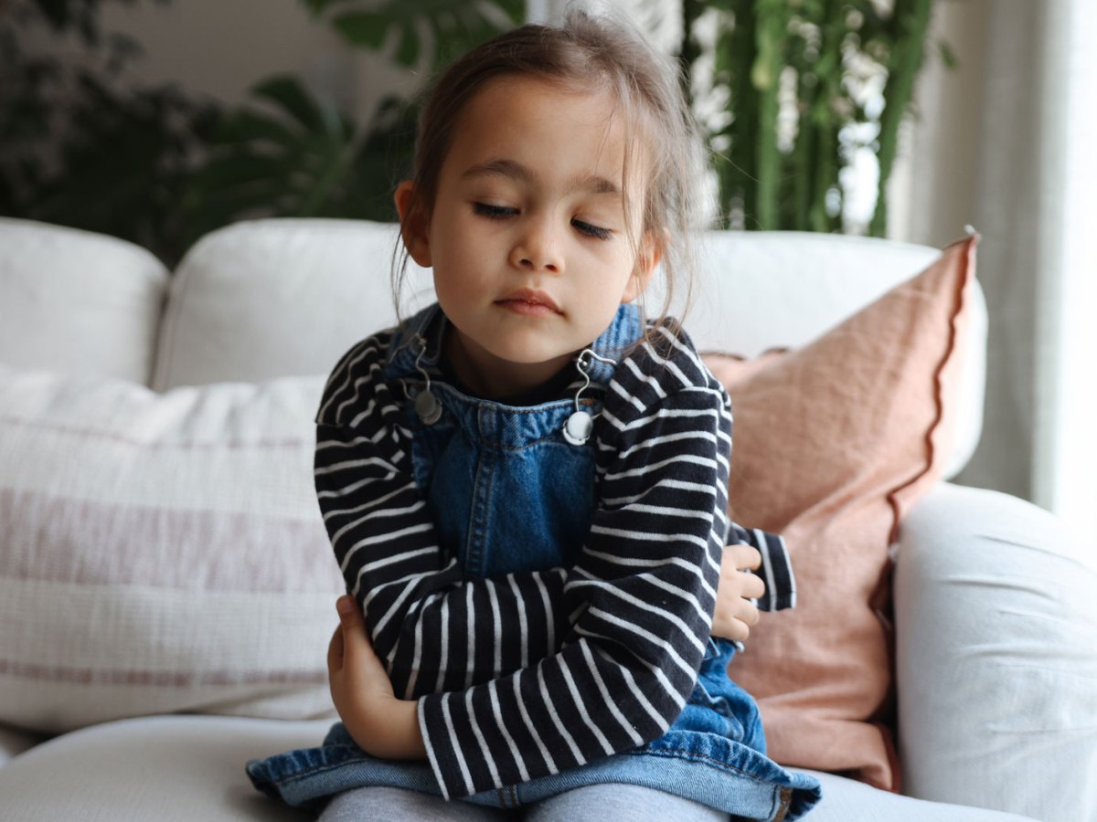 M&auml;dchen, ca. 4 Jahre alt, sitzt mit verschr&auml;nkten Armen auf dem Sofa und schaut traurig auf den Boden.