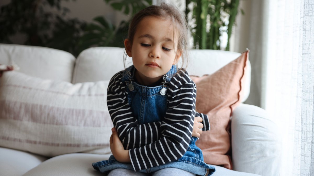 Mädchen, ca. 4 Jahre alt, sitzt mit verschränkten Armen auf dem Sofa und schaut traurig auf den Boden.