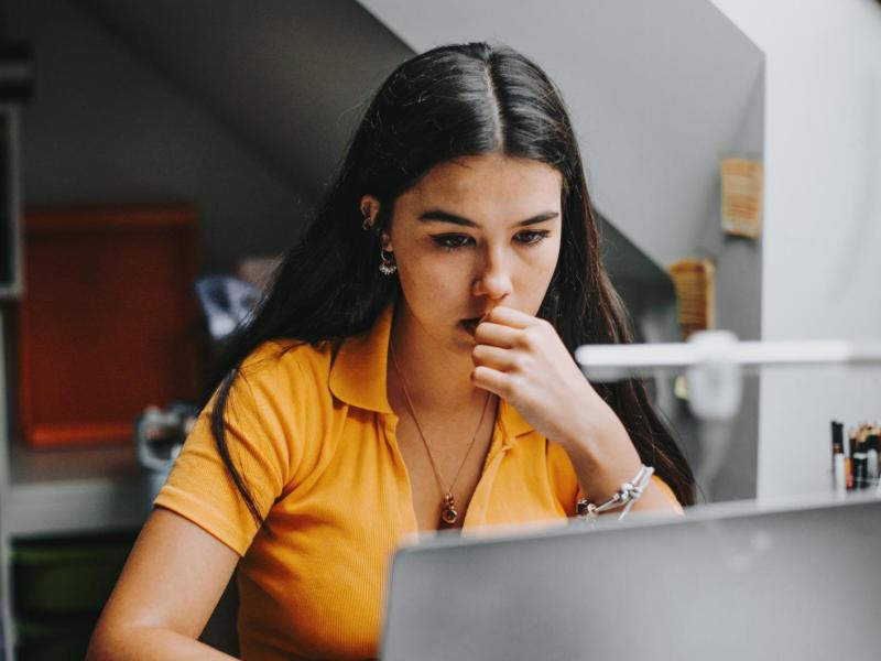 Junge Frau mit dunklen Haaren und senfgelben Shirt sitzt am Schreibtisch und schreibt eine Bewerbung an ihrem Laptop.