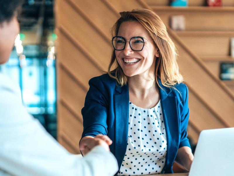 Frau sitzt auf der Arbeit an einem Tisch vor einem Laptop und gibt einer anderen Frau die Hand. Dabei l&auml;chelt sie stark.