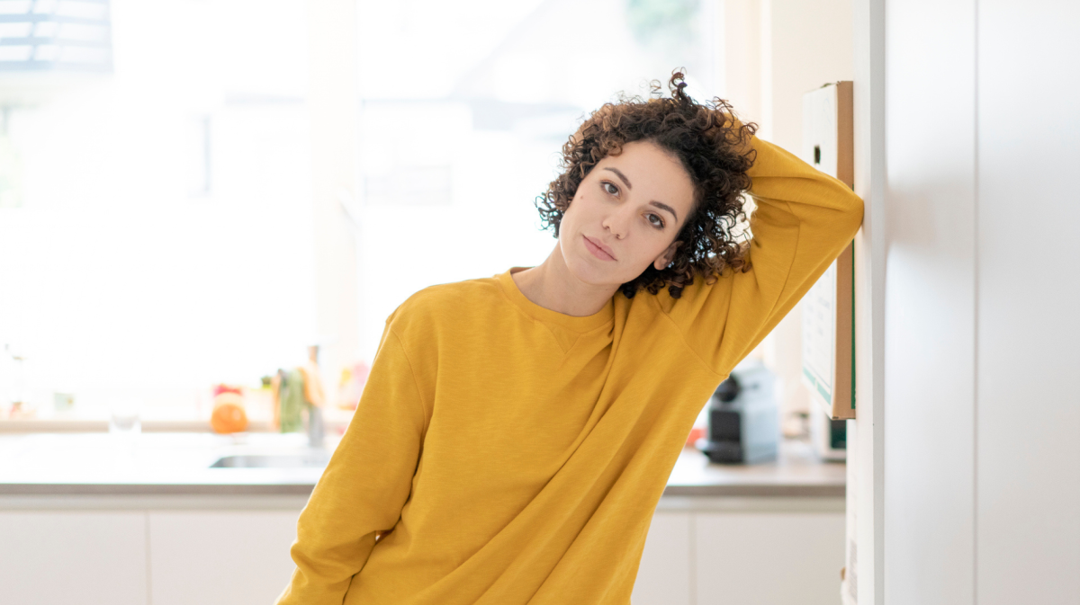 Frau mit dunklen Locken und einem gelben Pullover schaut ernst