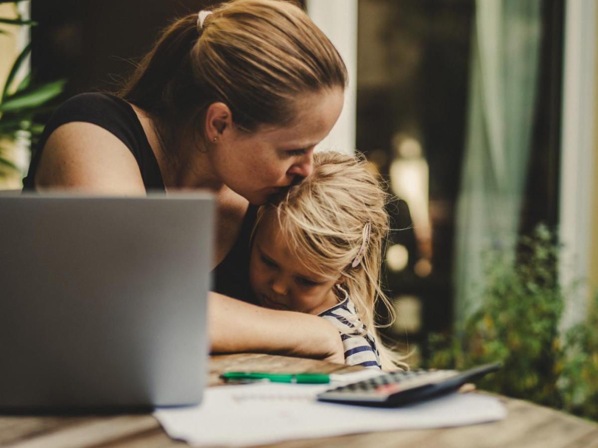 Frau arbeitet am Tisch und K&uuml;sst dabei ihre Tochter auf die Stirn, auf die sie neben der Arbeit aufpasst.
