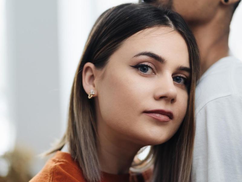 Frau guckt nachdenklich in die Kamera und lehnt sich dabei an die Brust ihres Mannes.