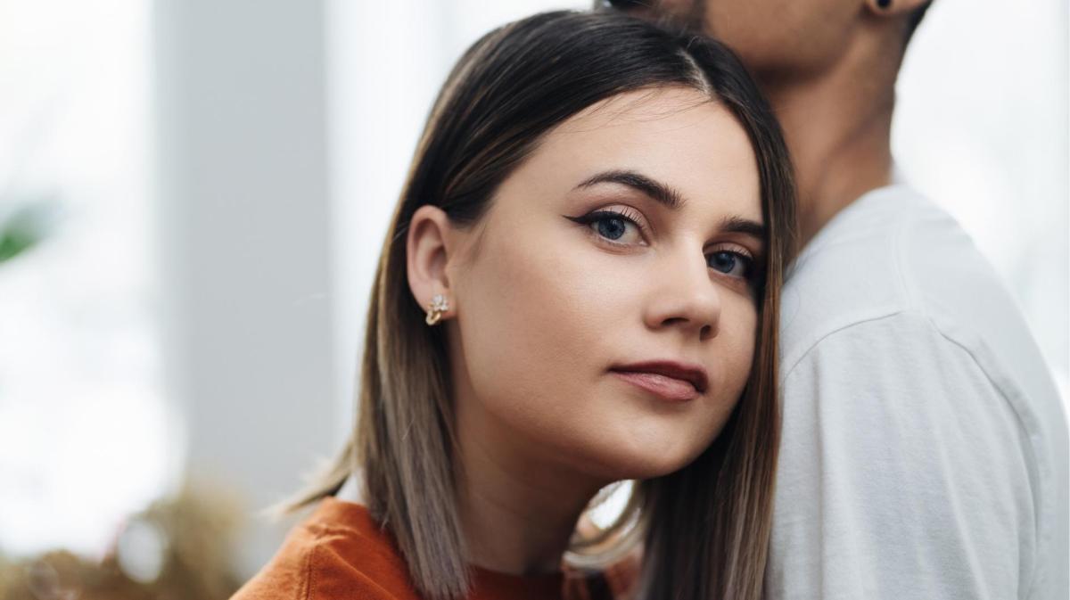 Frau guckt nachdenklich in die Kamera und lehnt sich dabei an die Brust ihres Mannes.