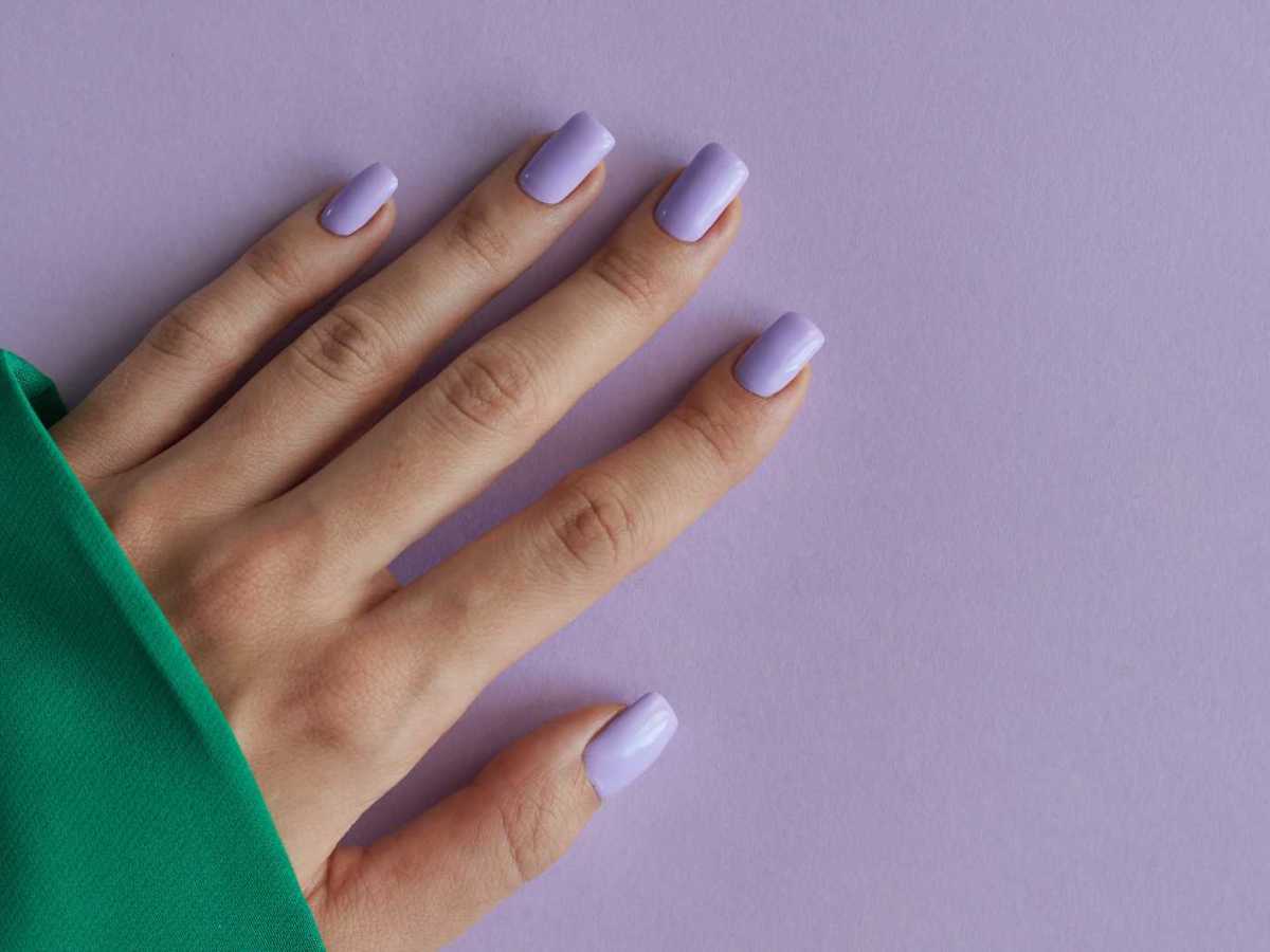 Hand mit lila lackierten Fingernägeln und einem grünen Oberteil vor einem fliederfarbenen Hintergrund