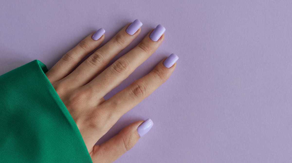 Hand mit lila lackierten Fingernägeln und einem grünen Oberteil vor einem fliederfarbenen Hintergrund