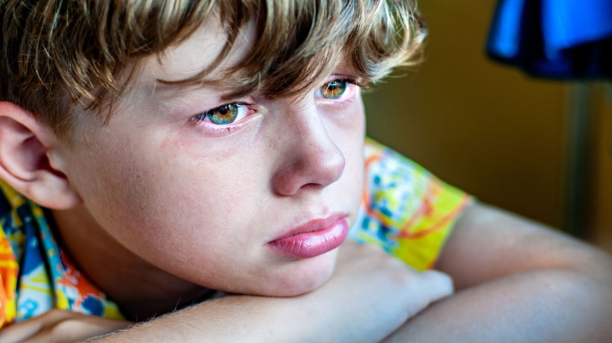 Profil eines traurigen Jungen, der den Kopf auf den auf einem Tisch liegenden verschränkten Armen abgelegt hat.
