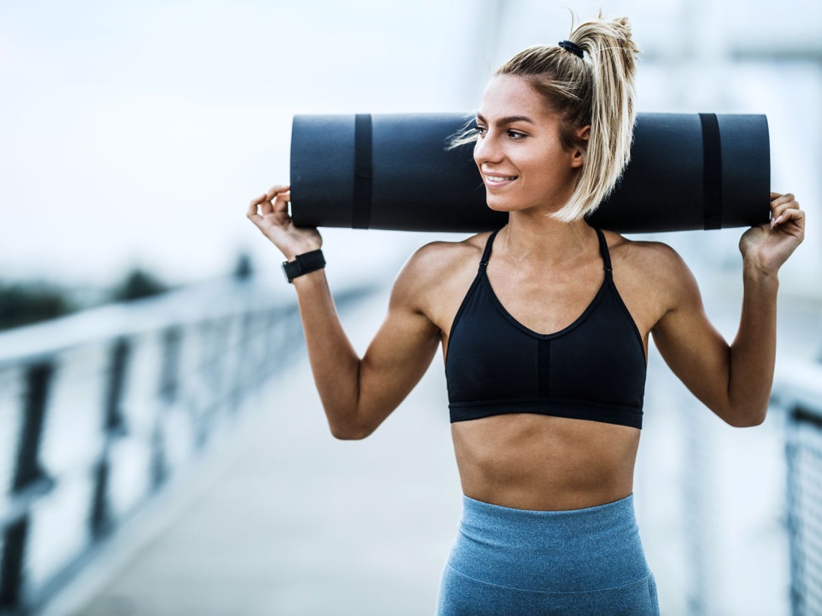 Sportliche Frau mit trainiertem K&ouml;rper tr&auml;gt Sportmatte &uuml;ber den Schultern