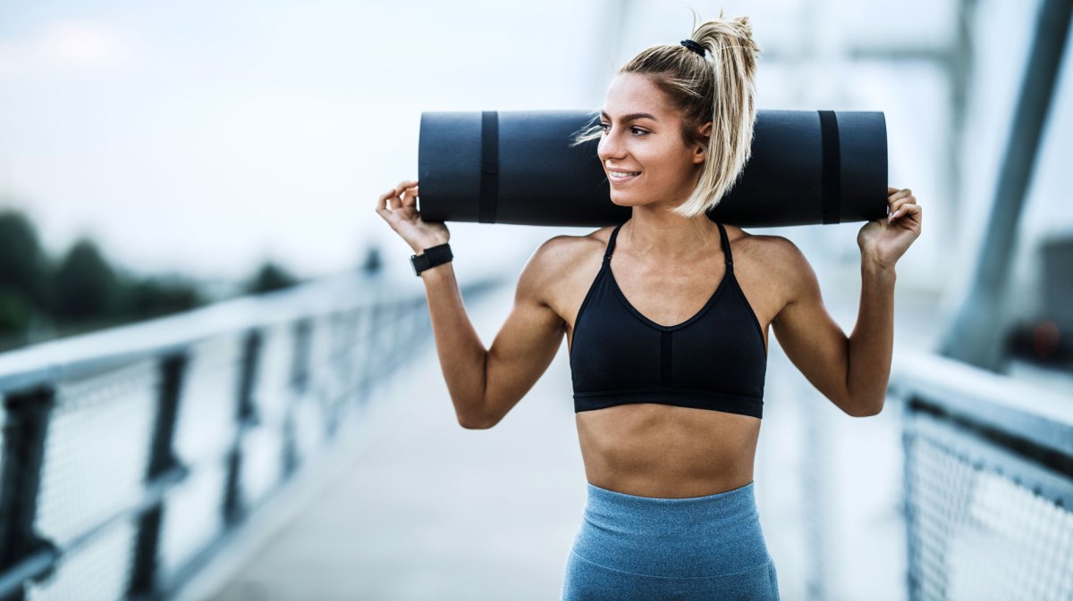 Sportliche Frau mit trainiertem Körper trägt Sportmatte über den Schultern