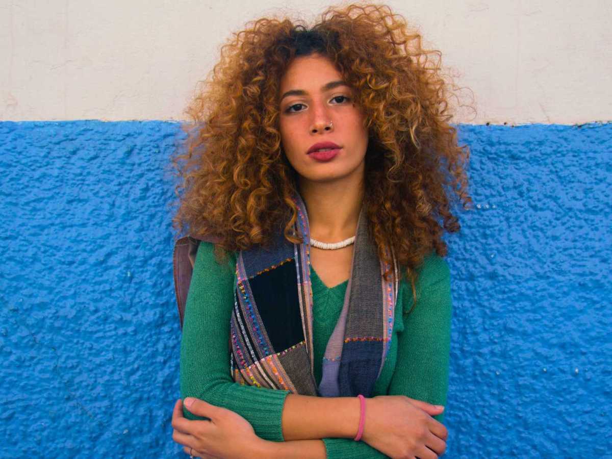 Junge Frau mit Locken steht vor einer blau-wei&szlig;en Wand und schaut in die Kamera.