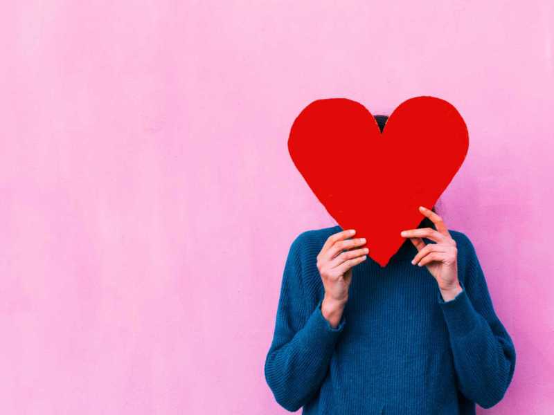 Frau steht vor einer pinken Wand und versteckt ihr Gesicht hinter einem großen Herz.