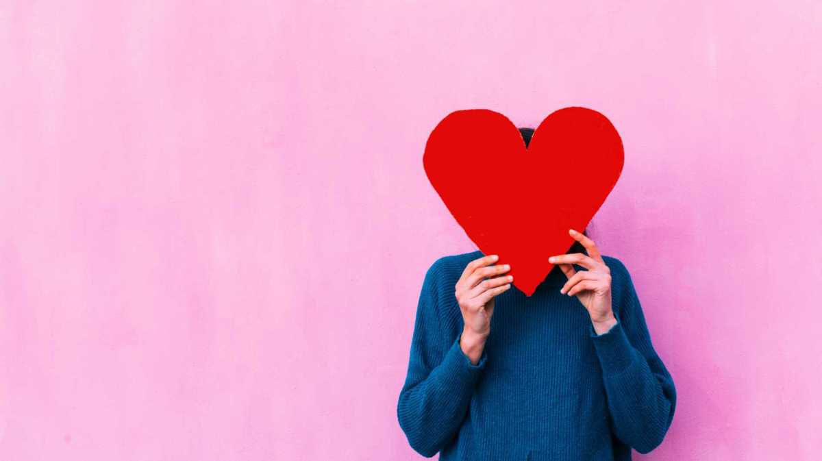 Frau steht vor einer pinken Wand und versteckt ihr Gesicht hinter einem großen Herz.