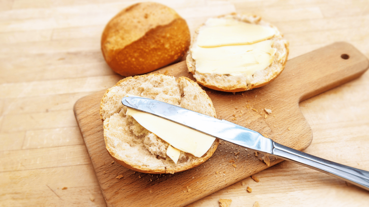 Aufgeschnittenes Brötchen auf einem Holzbrettchen, mit Butter bestrichen.