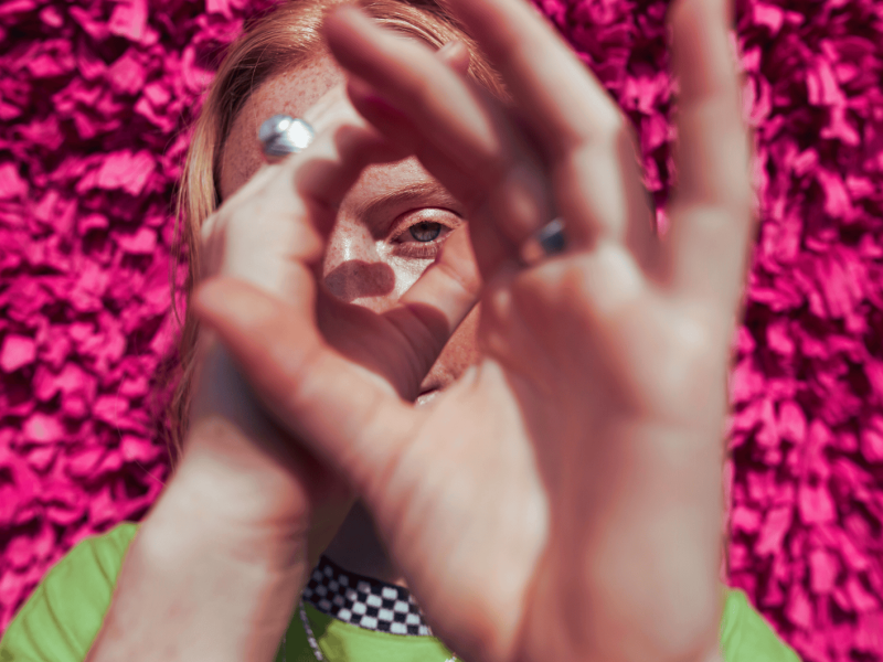 Frau mit Sommersprossen steht vor einem pinken Hintergrund und schaut durch ihre Hände, die ein Fernrohr bilden.
