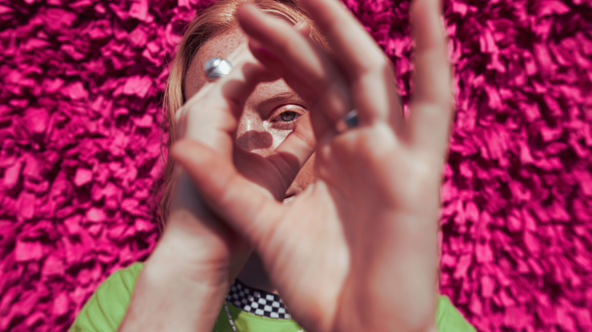 Frau mit Sommersprossen steht vor einem pinken Hintergrund und schaut durch ihre Hände, die ein Fernrohr bilden.