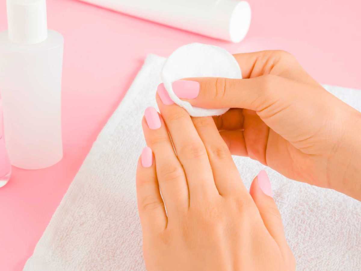 Junge Frau entfernt rosa Nagellack mit einem wei&szlig;en Wattepad auf einem Handtuch auf einem hellrosa Tischhintergrund. Nahaufnahme.