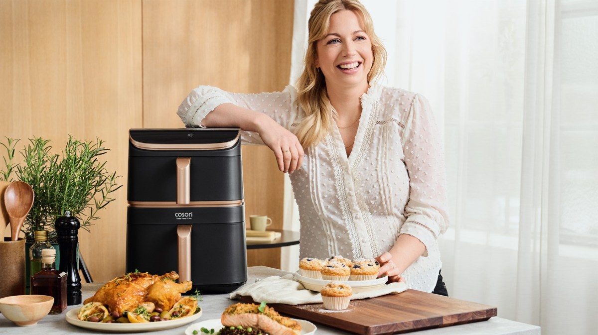 Junge Frau mit halboffenem Haar präsentiert fröhlich Muffins neben moderner Heißluftfritteuse und frisch gekochtem Essen in heller Küche.