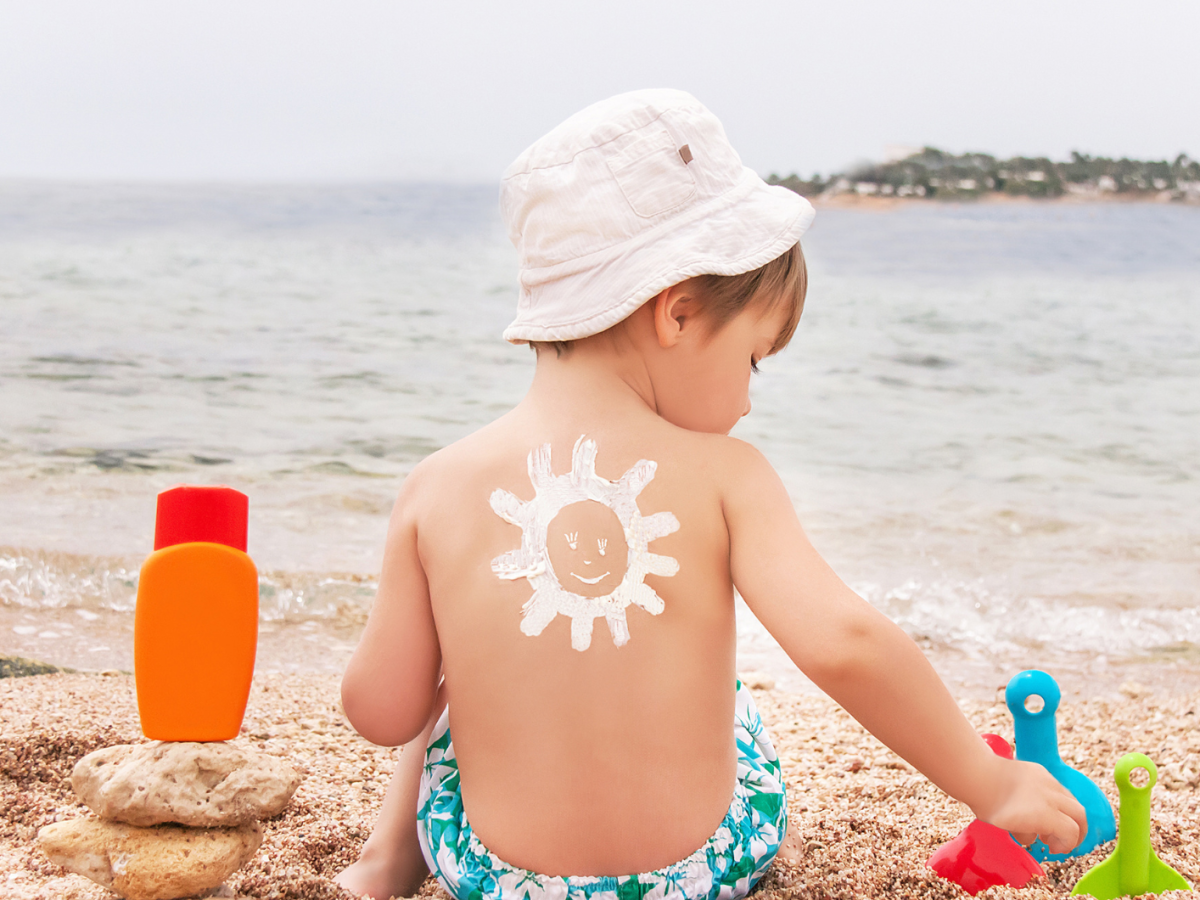 Kleiner Junge sitzt in Badekleidung am Strand und hat auf dem R&uuml;cken eine Sonne aus Sonnencreme gemalt