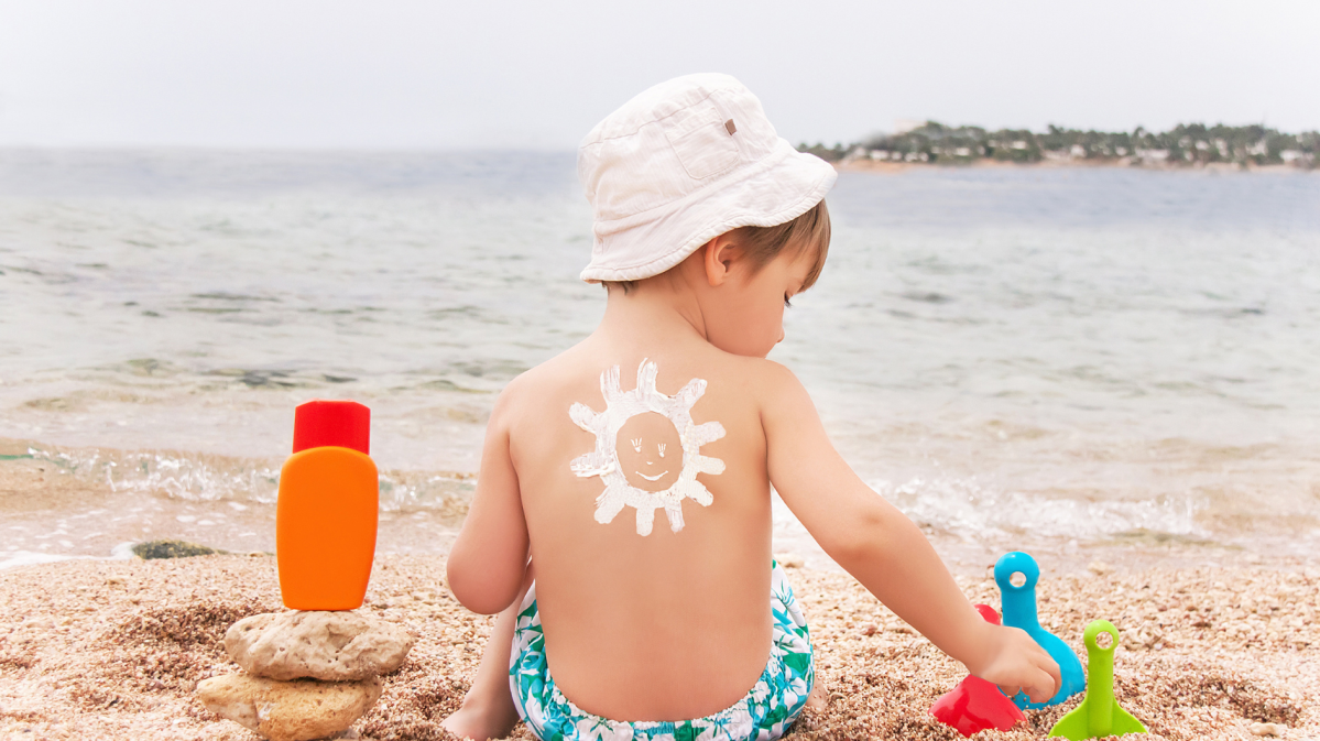 Kleiner Junge sitzt in Badekleidung am Strand und hat auf dem Rücken eine Sonne aus Sonnencreme gemalt