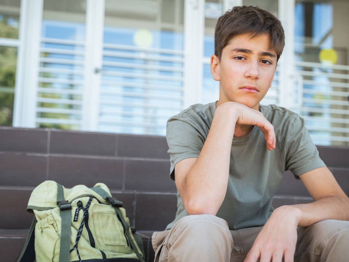 Junge im Teenageralter sitzt traurig auf den Treppenstufen vor seiner Schule, neben ihm ein Rucksack