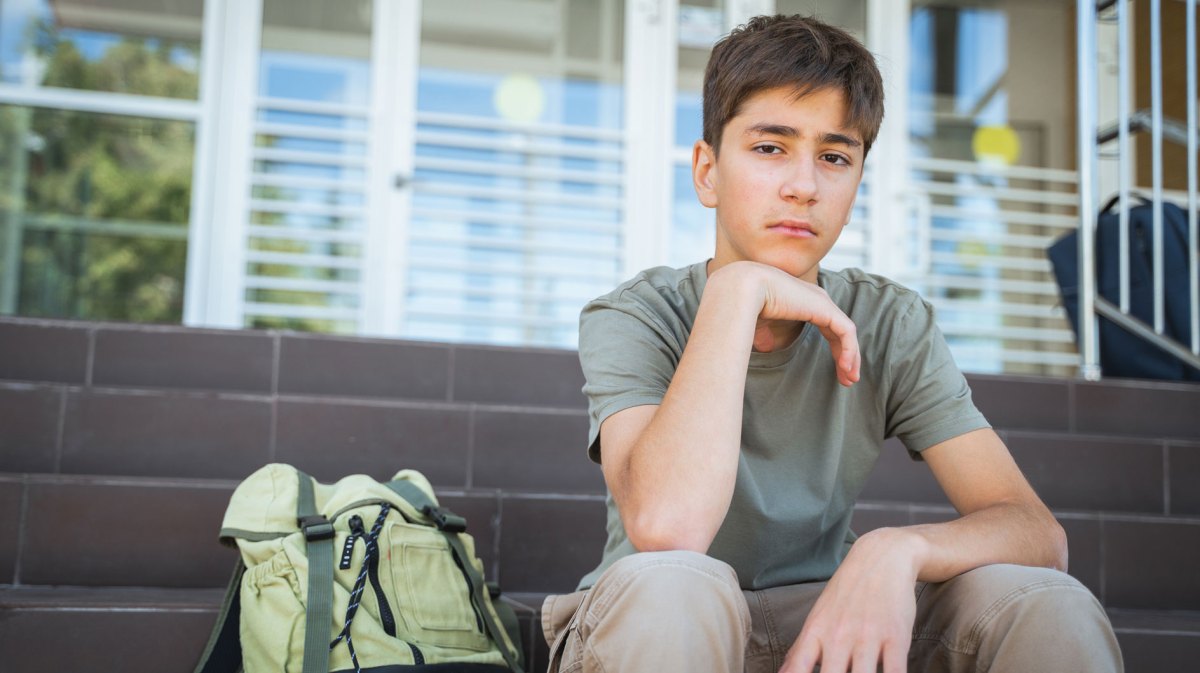 Junge im Teenageralter sitzt traurig auf den Treppenstufen vor seiner Schule, neben ihm ein Rucksack