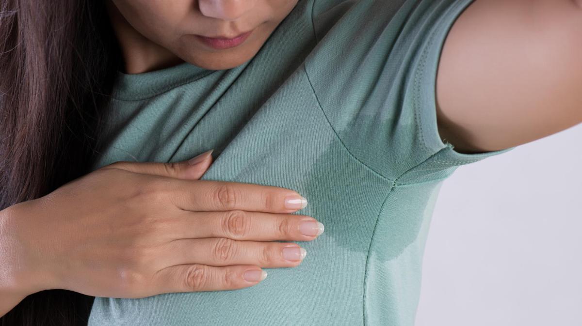 Frau guckt unter ihre Achsel. Sie hat einen Schweißfleck, der auf dem grünen Shirt deutlich sichtbar ist.