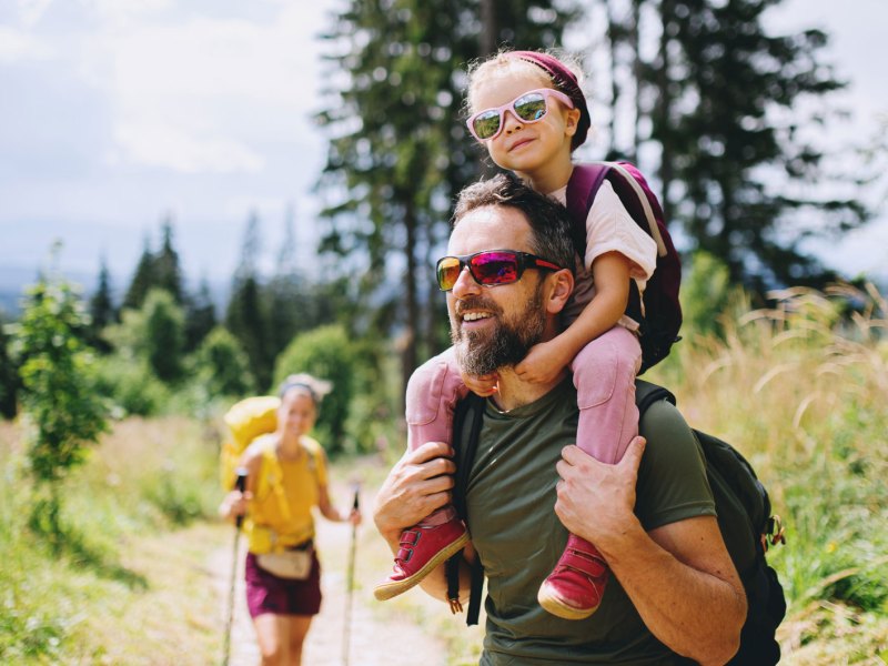 Eltern gehen mit ihrem Kind wandern. Der Vater trägt die Tochter auf den Schultern.