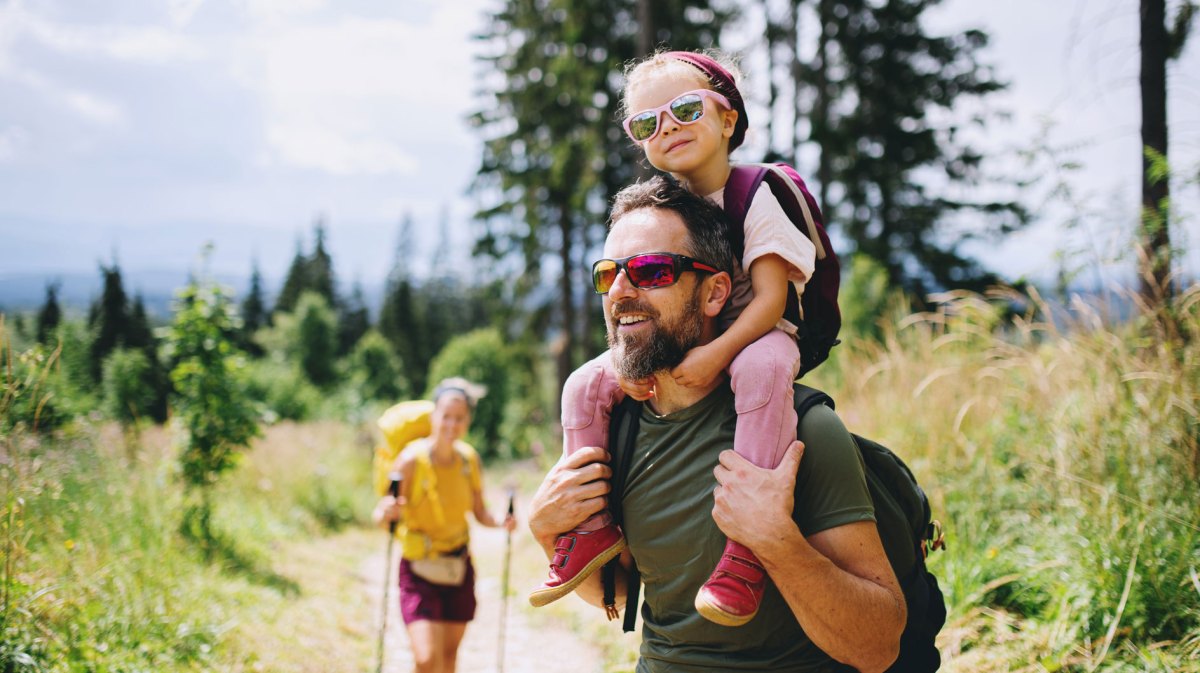 Eltern gehen mit ihrem Kind wandern. Der Vater trägt die Tochter auf den Schultern.