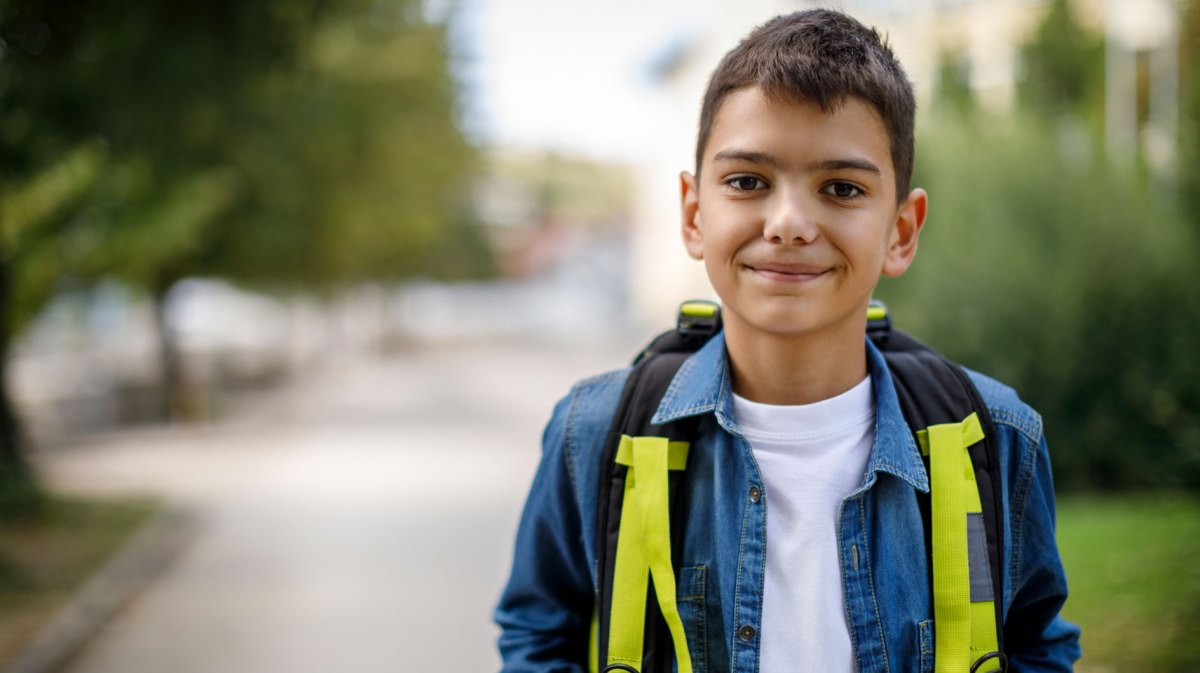 Porträt eines Jungen, ca. 10 Jahre alt, steht mit einem Rucksack auf der Straße und schaut freundlich in die Kamera.
