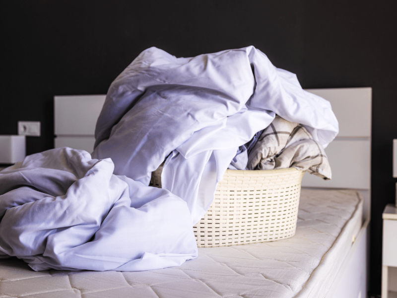 Wäschekorb mit Bettwäsche steht auf einem abgezogenen Bett.