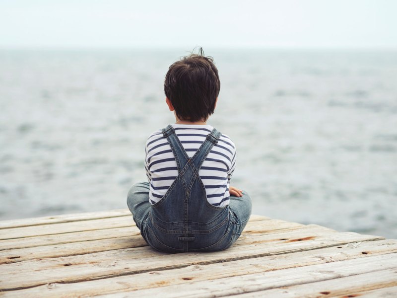 Kind sitzt mit dem Rücken zur Kamera auf einem Steg und schaut aufs Meer.