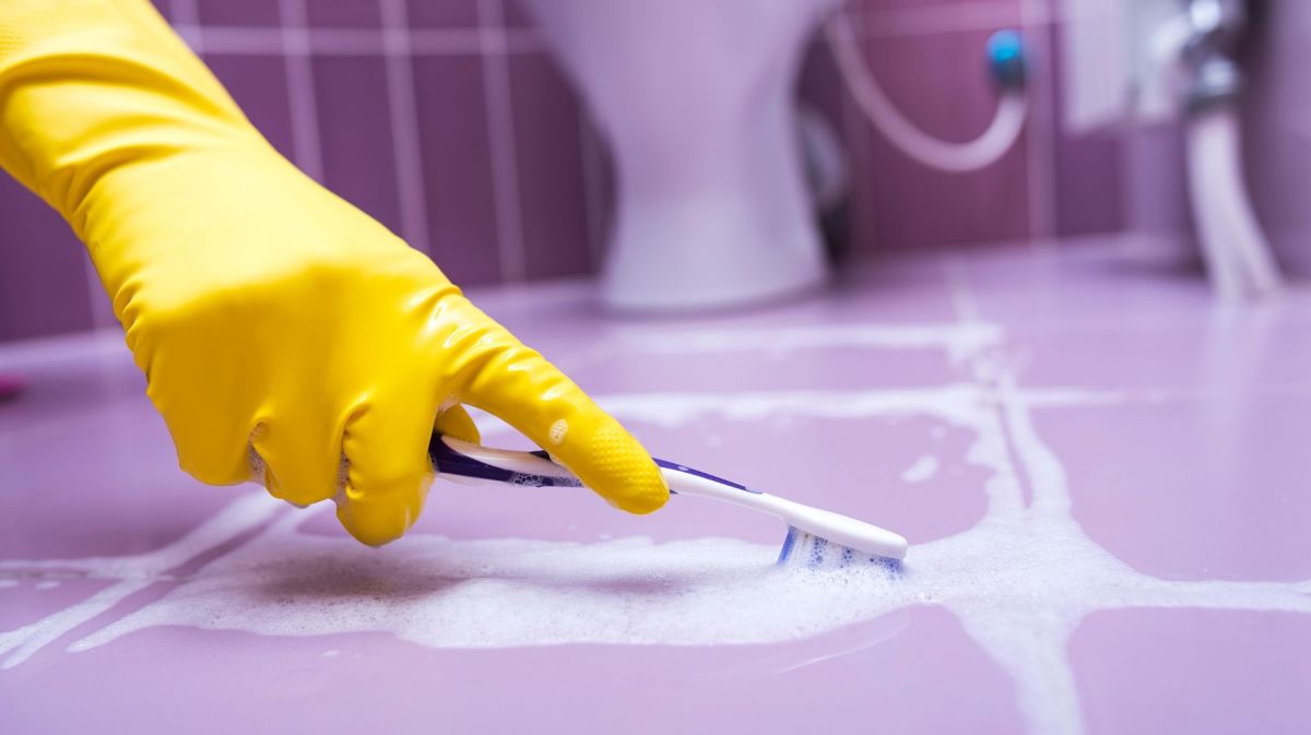 Hand mit gelbem Gummihandschuh putzt mit Zahnbürste Silikonfugen zwischen lila Fugen